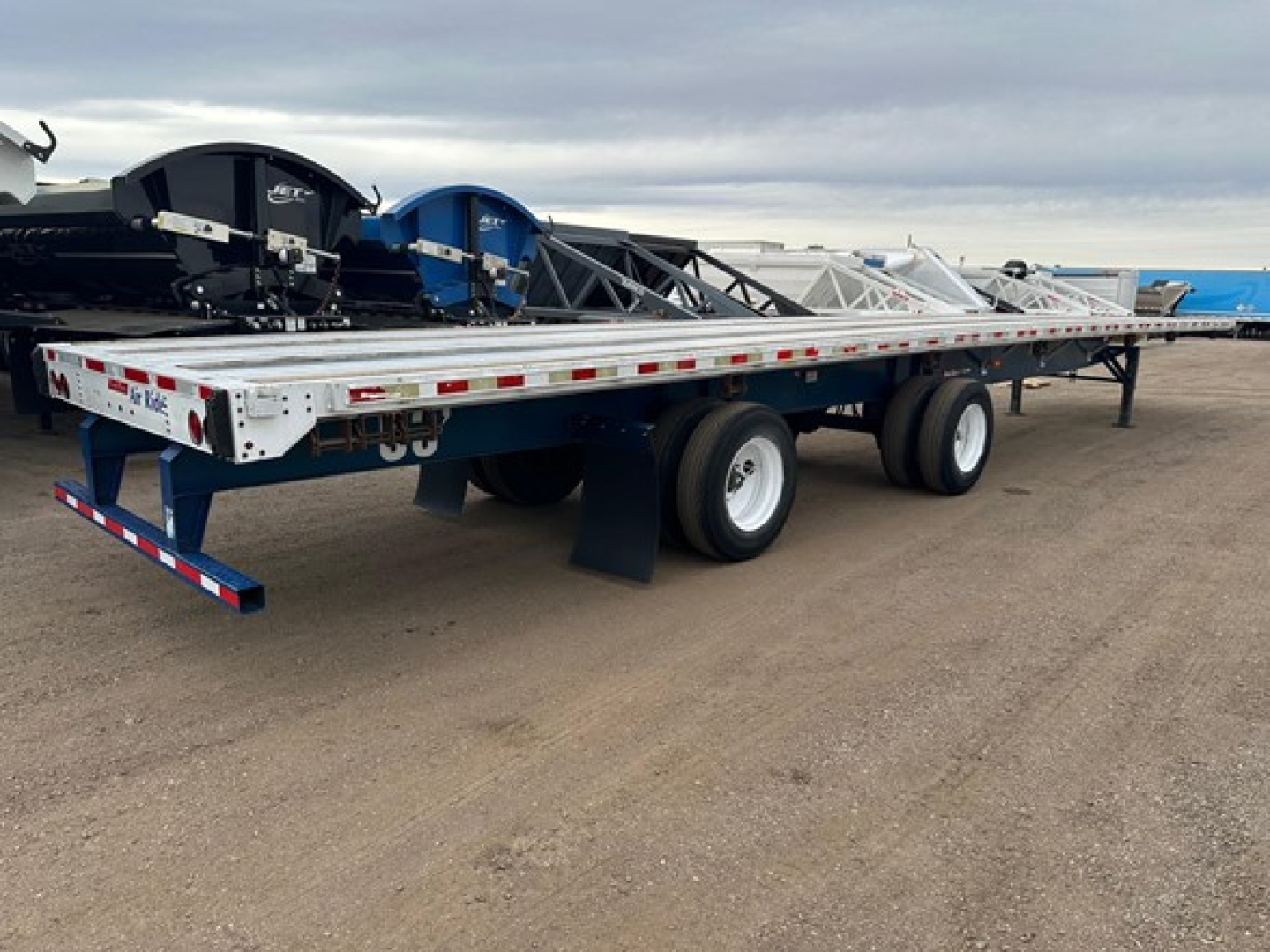 Flatbed trailer with dual rear axles and white frame, viewed from side, carrying equipment and coil rollers.