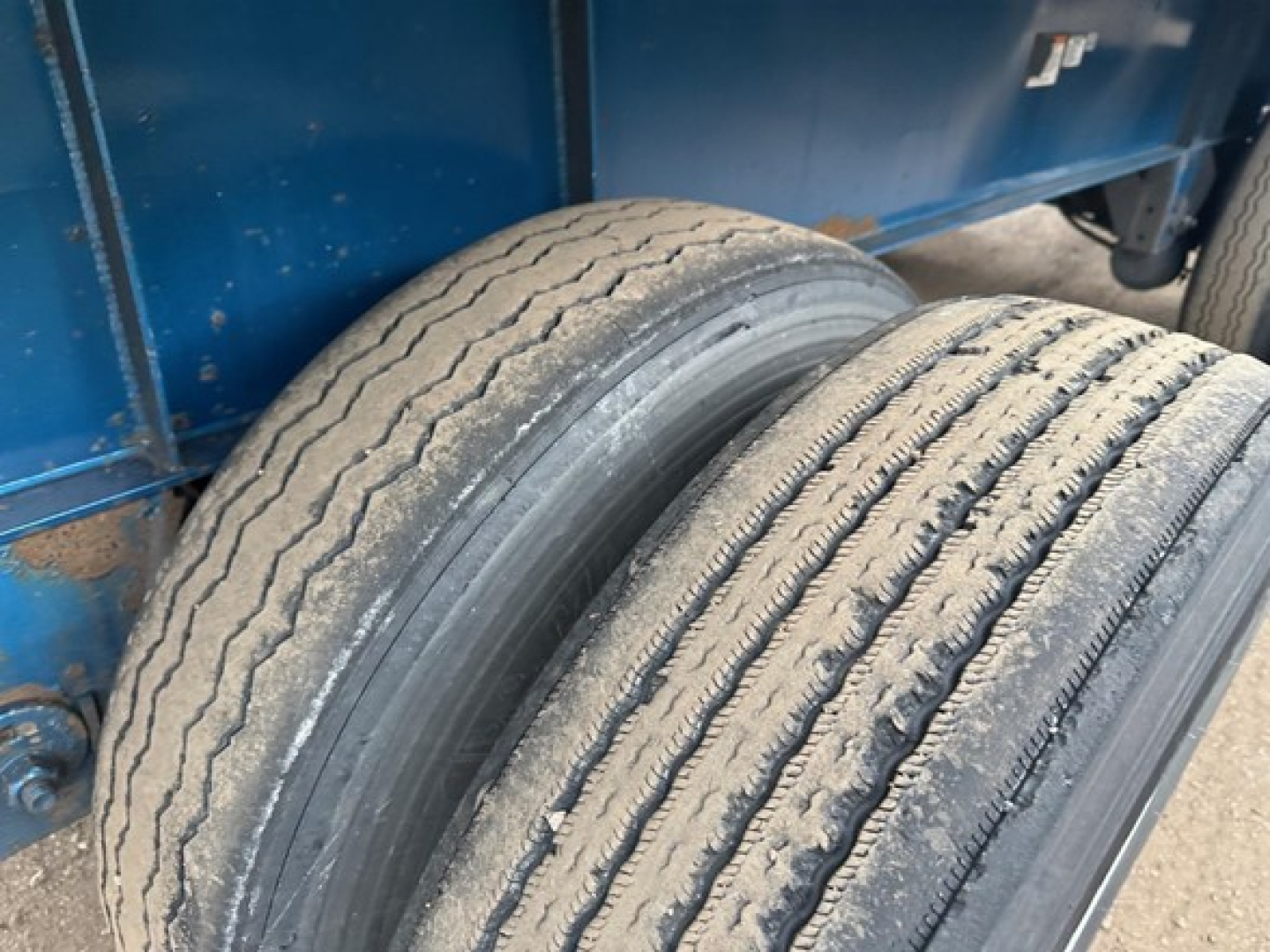 Trailer axle with dual tires showing tread wear and blue trailer body in background.