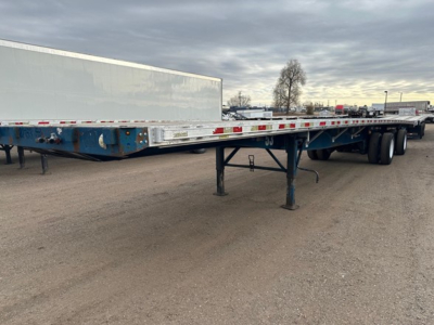 Side view of a flatbed trailer with blue frame and white gooseneck storage box, extended landing gear down, dual rear axles, in a gravel lot.