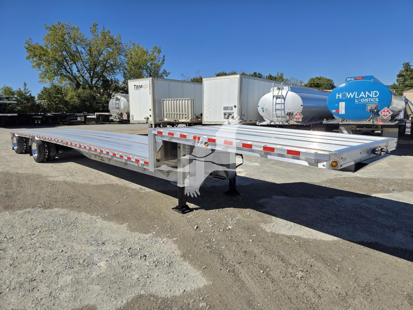 Trailer in a lot showing a tandem-axle dropdeck flatbed with aluminum deck, under-mounted landing gear, and reflective safety rails; Howland Logistics tankers and storage units visible in background.