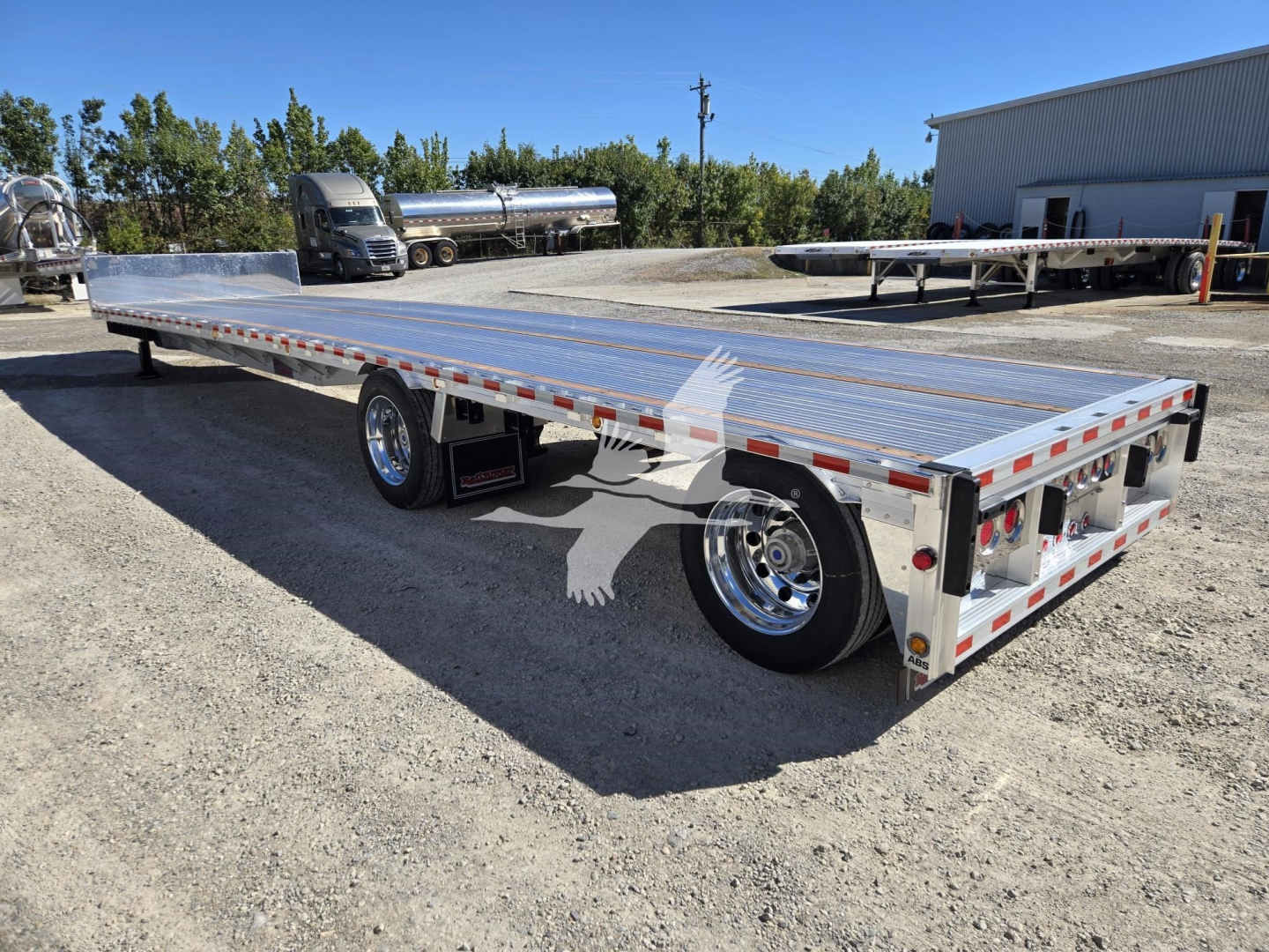 Trailer showing a long aluminum dropdeck flatbed with rear ramps, dual rear axles, chrome wheels, and reflective side markings.