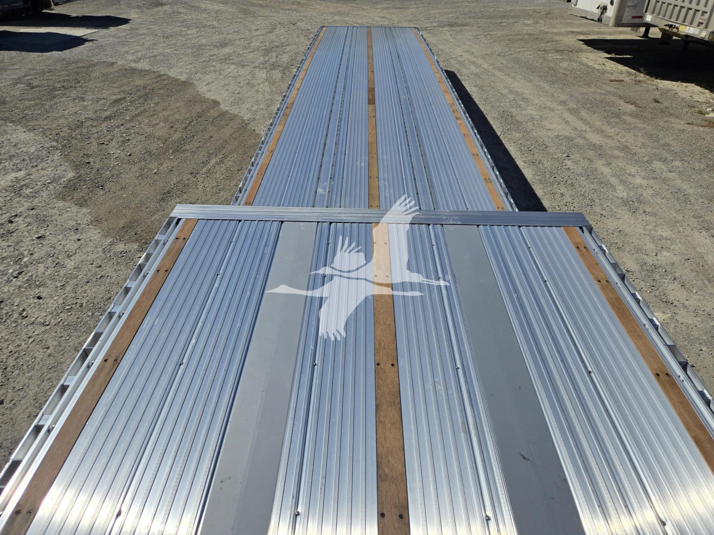 Trailer deck showing aluminum dropdeck with long unpainted rails and a white bird logo; VIN tag not visible.