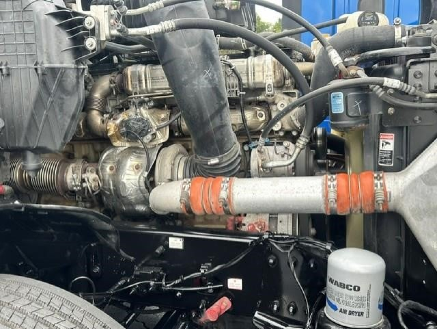 Truck engine bay with various hoses, belts, and a white cylindrical filter labeled WABCO; orange coolant hose; black metal frame and components visible.
