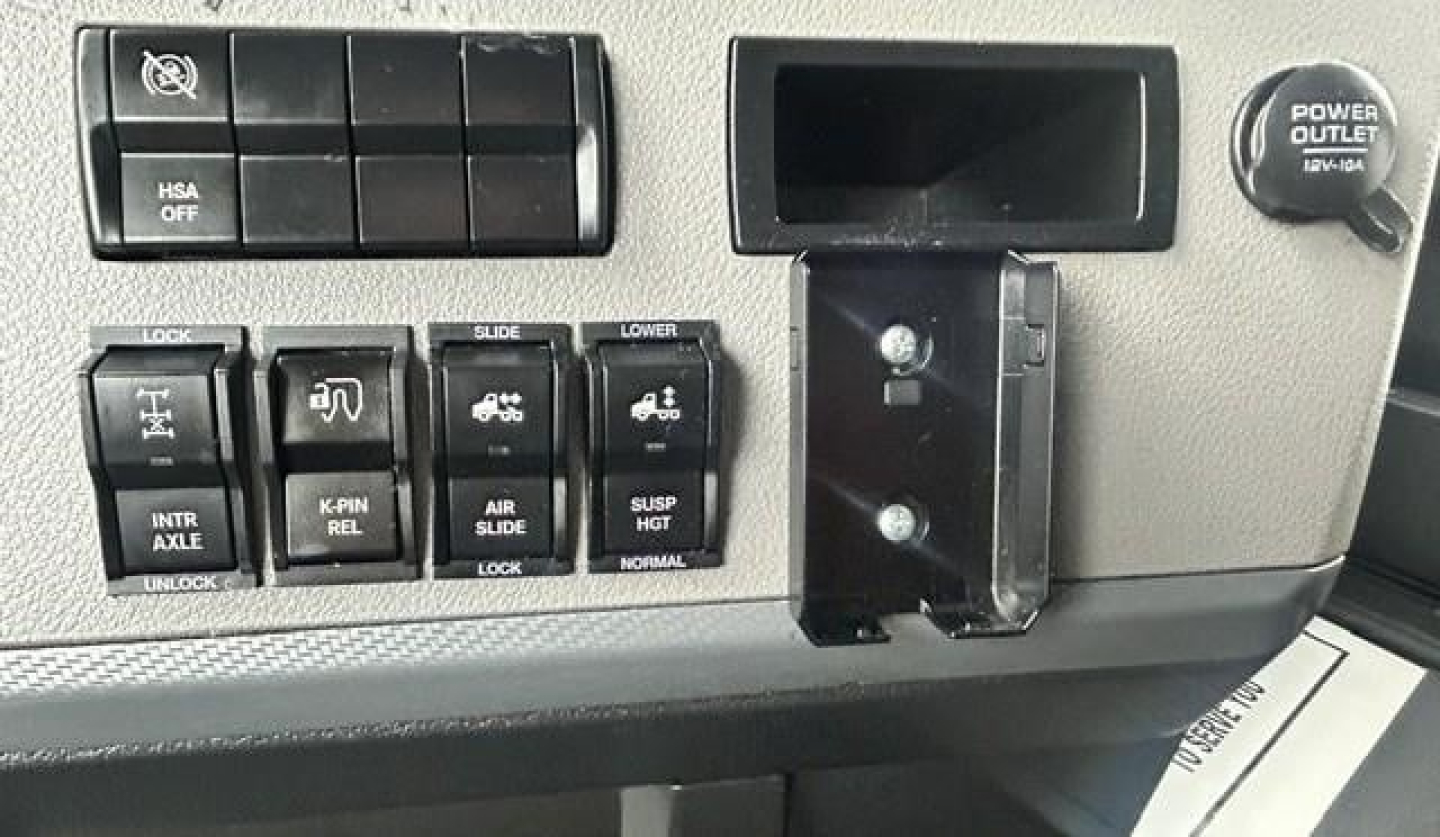 Cabin control panel with multiple switches and a power outlet on Freightliner Cascadia dash, showing various labeled buttons and a small display pocket.