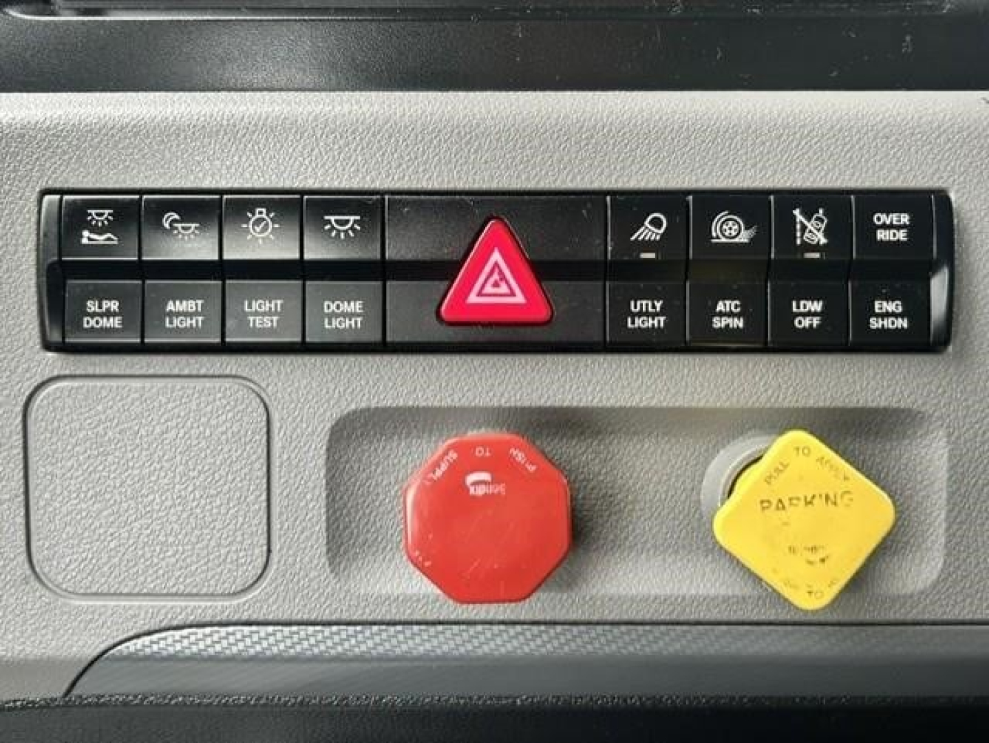 Dashboard panel of a Freightliner Cascadia showing row of buttons including hazard light, light controls, and two colored knobs (red and yellow) with text on them.