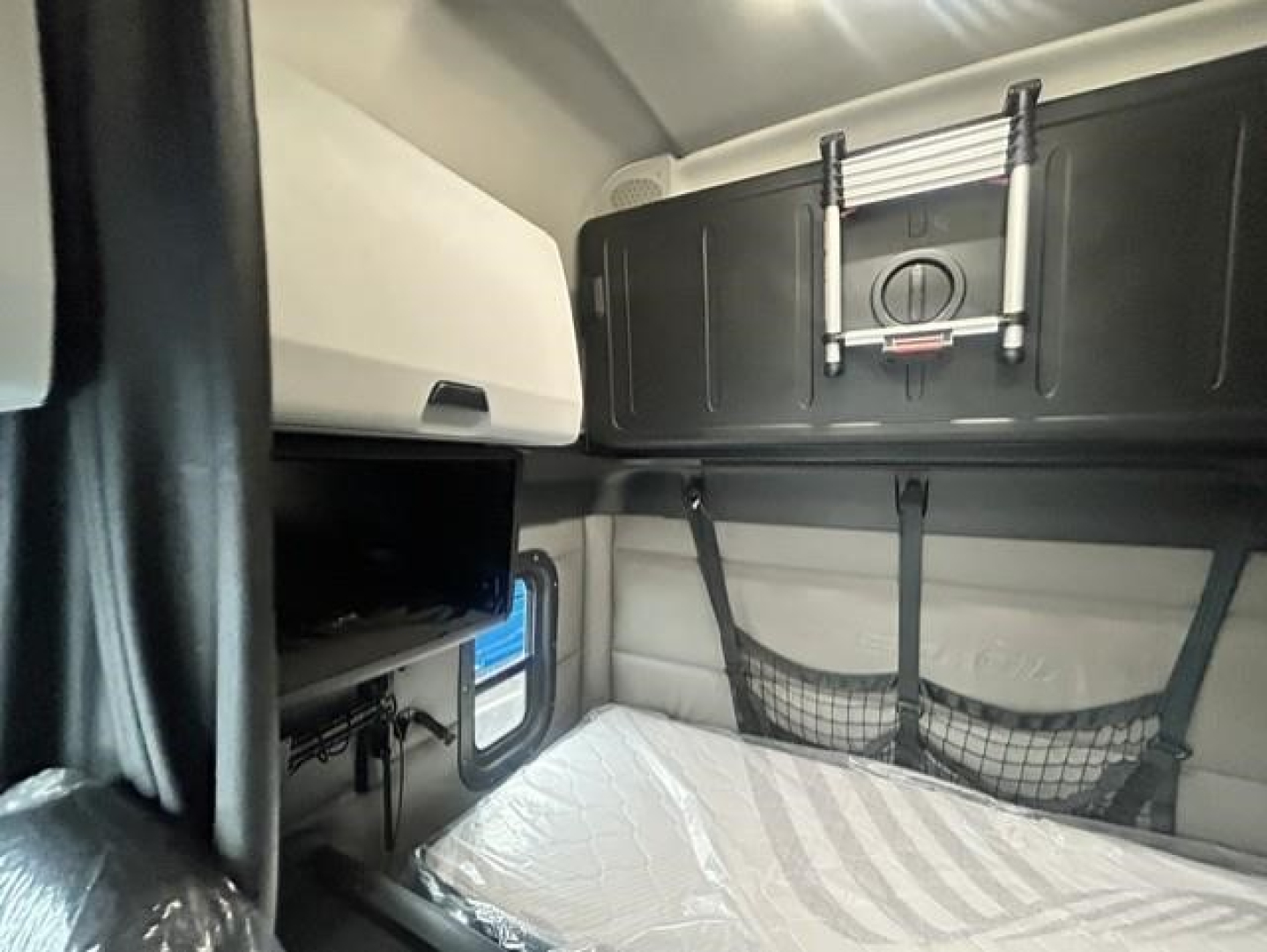 Interior sleeper cab of a Freightliner Cascadia showing bunk bed area with storage cabinet and seat; compact layout.