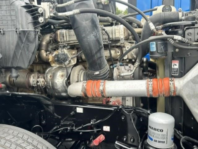 Truck engine bay with various hoses, belts, and a white cylindrical filter labeled WABCO; orange coolant hose; black metal frame and components visible.