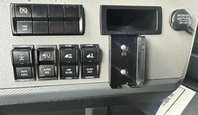 Cabin control panel with multiple switches and a power outlet on Freightliner Cascadia dash, showing various labeled buttons and a small display pocket.