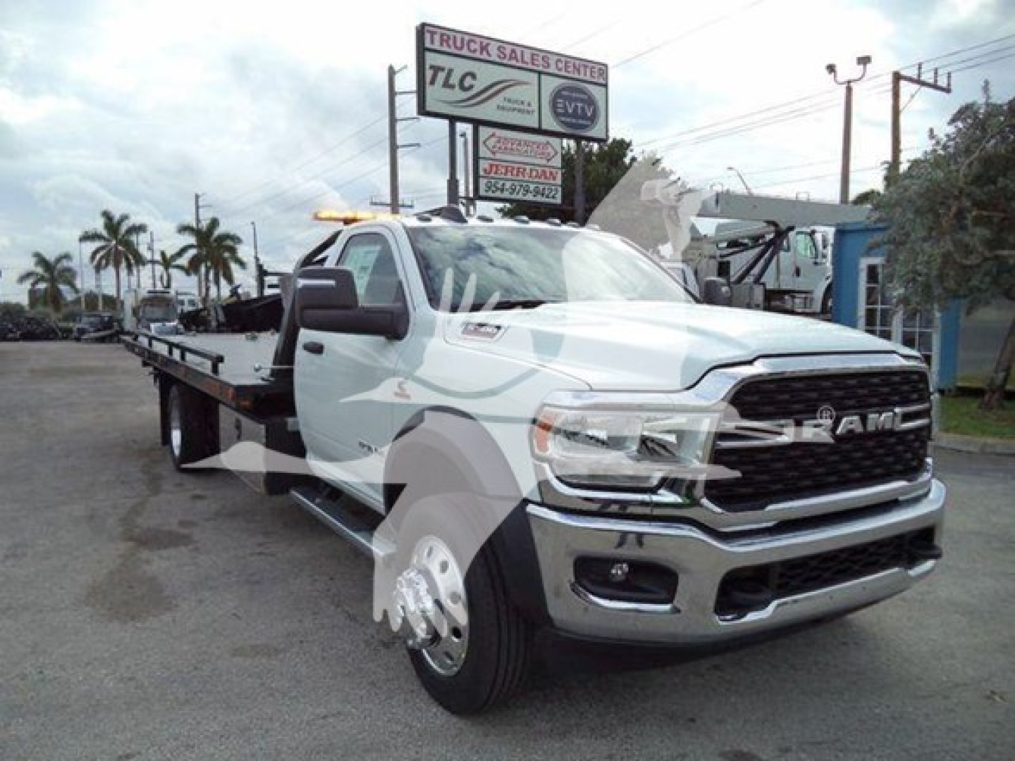 Flatbed tow truck with white cab and black bed, RAM grille, large side mirrors, dealership sign in background. Visible text includes TLC Truck Sales Center on sign; no other legible model details visible on vehicle.