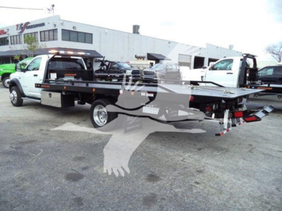 Ram 5500 rollback tow truck with extended flatbed in a dealership lot, cab white, equipment on bed, multiple trucks in background.