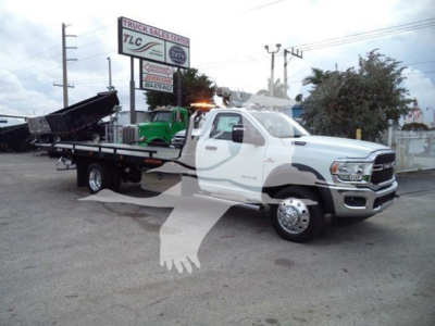 Ram 5500 tow truck in a lot with a rollback bed; front grille with RAM logo, white cab, and equipment on bed; dealership sign visible in background; some text on sign and vehicles.