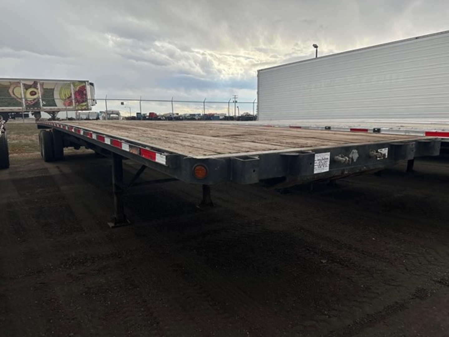 Flatbed trailer deck with long metal frame, Fontaine Velocity-style trailer, visible reflective tape along edge, landing gear legs under the front, white sticker near the side.