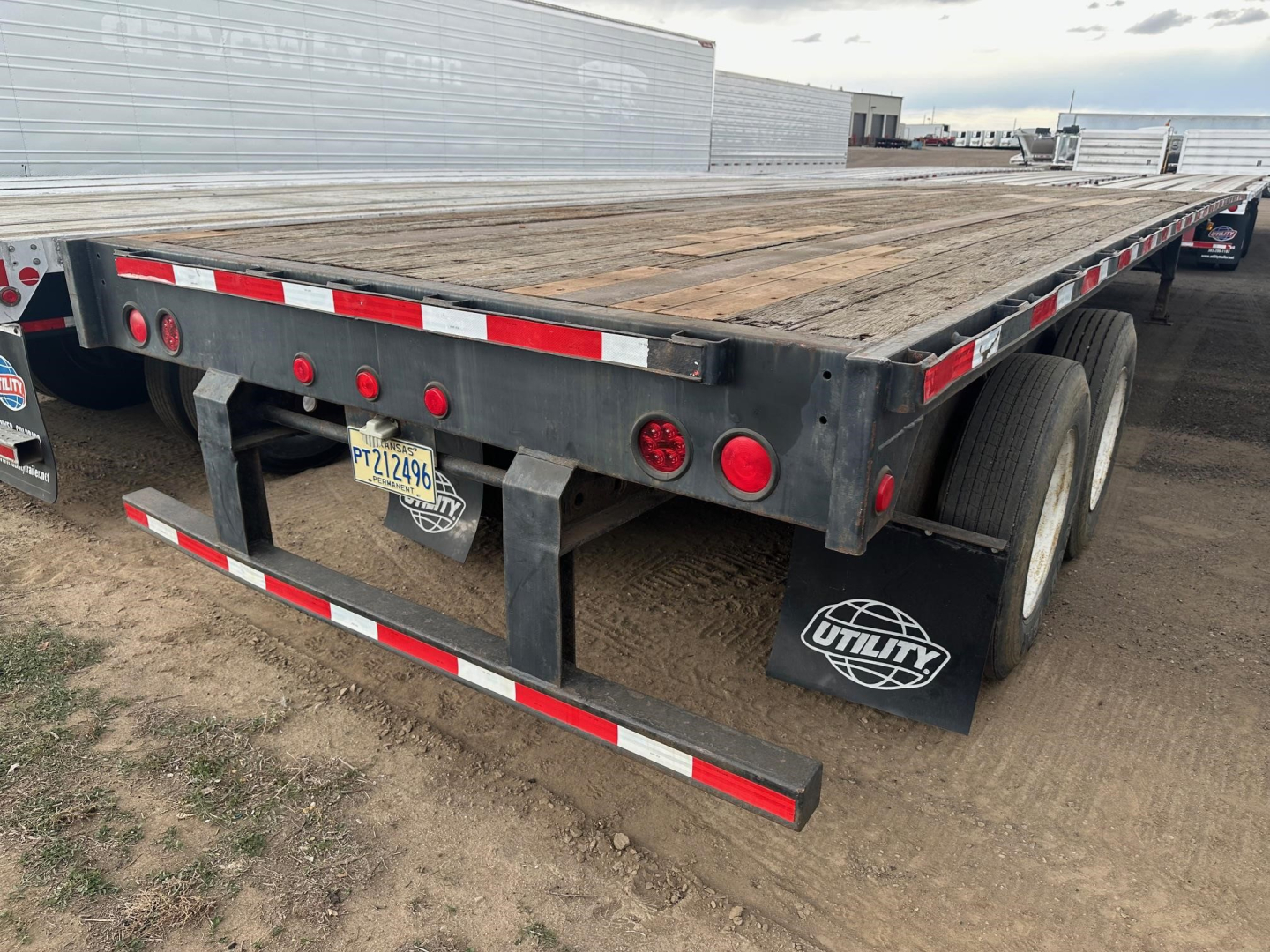 Flatbed trailer rear view showing wooden deck, dual rear axles, mudflaps with Utility brand, and light assembly with reflectors.