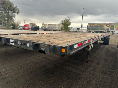 Flatbed trailer in service showing wooden deck, side reflectors, and dual rear axles with a drop tail.