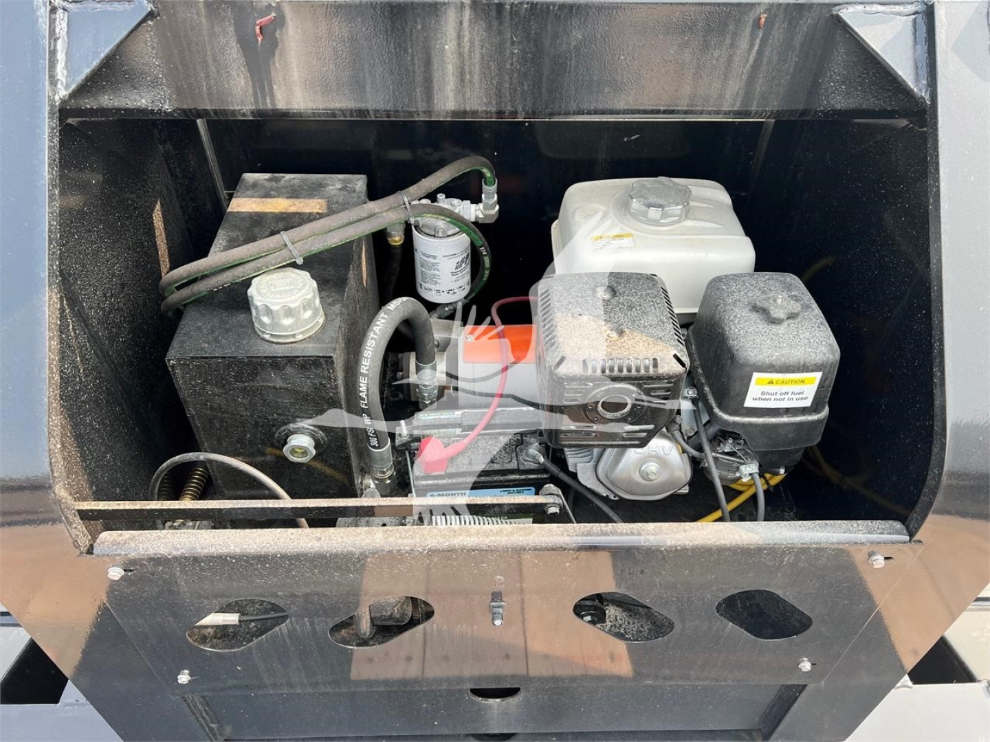 Trailer engine bay showing compact 4-stroke engine with white coolant reservoir, black hydraulic tank, and wiring in a lowboy trailer compartment.