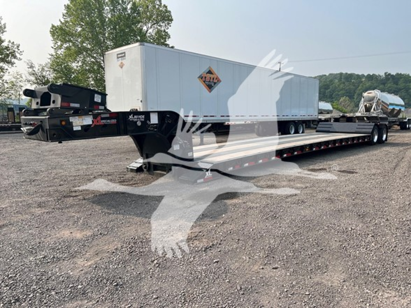 Lowboy trailer with a white box trailer on a long black flatbed; trailer hitch area shows branding and a spare tire rack; distant hills and gravel lot.