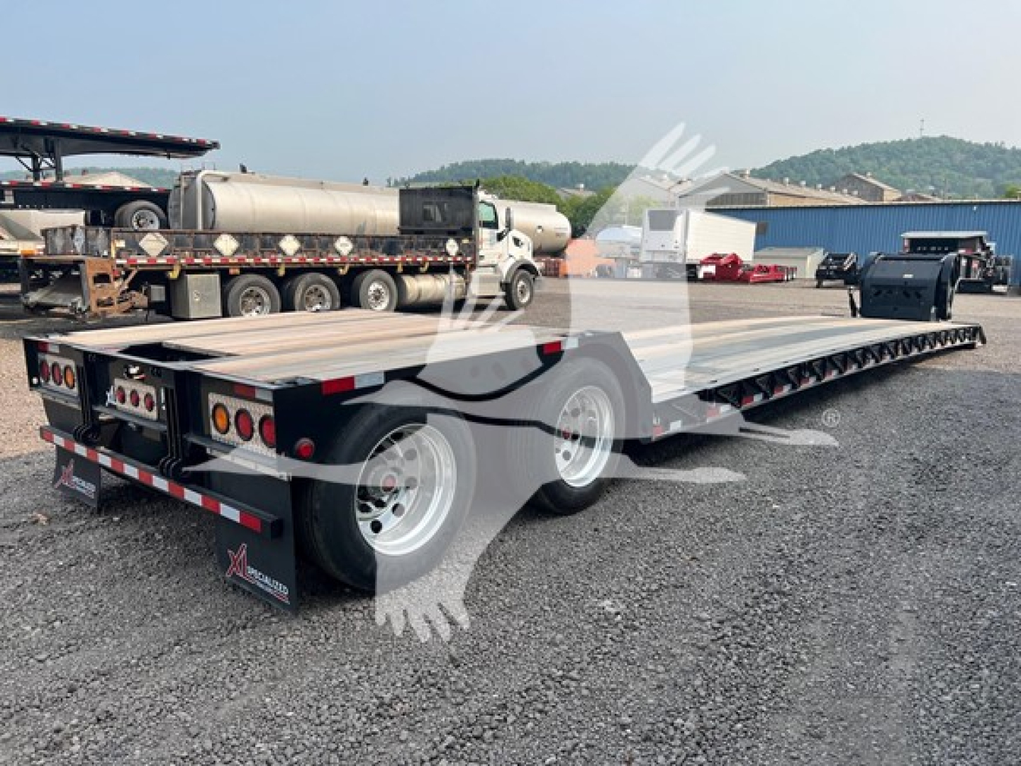 XL Specialized XL80HD E lowboy trailer showing flatbed with dual rear axles and taillights, silver aluminum deck, black frame, in a gravel lot.
