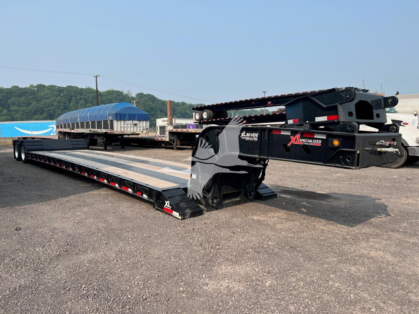 XL Specialized XL80HD trailer in lowboy configuration with extended deck and gooseneck, black frame, blue tarps in background, equipment on site.