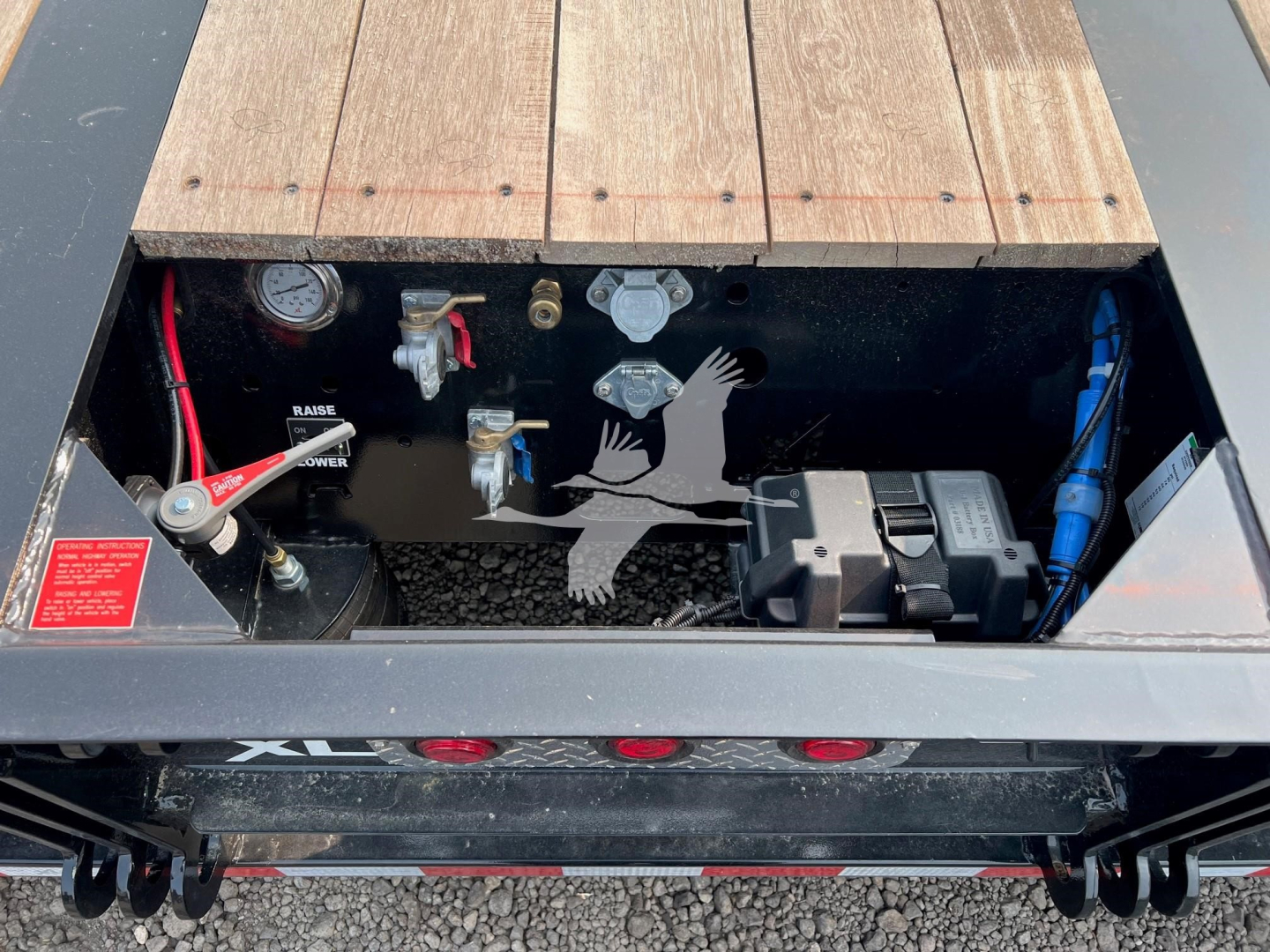 Open storage compartment of XL Specialized XL80HDE trailer showing battery, hydraulic reservoir, gauges, and wiring; label text and controls visible inside toolbox.