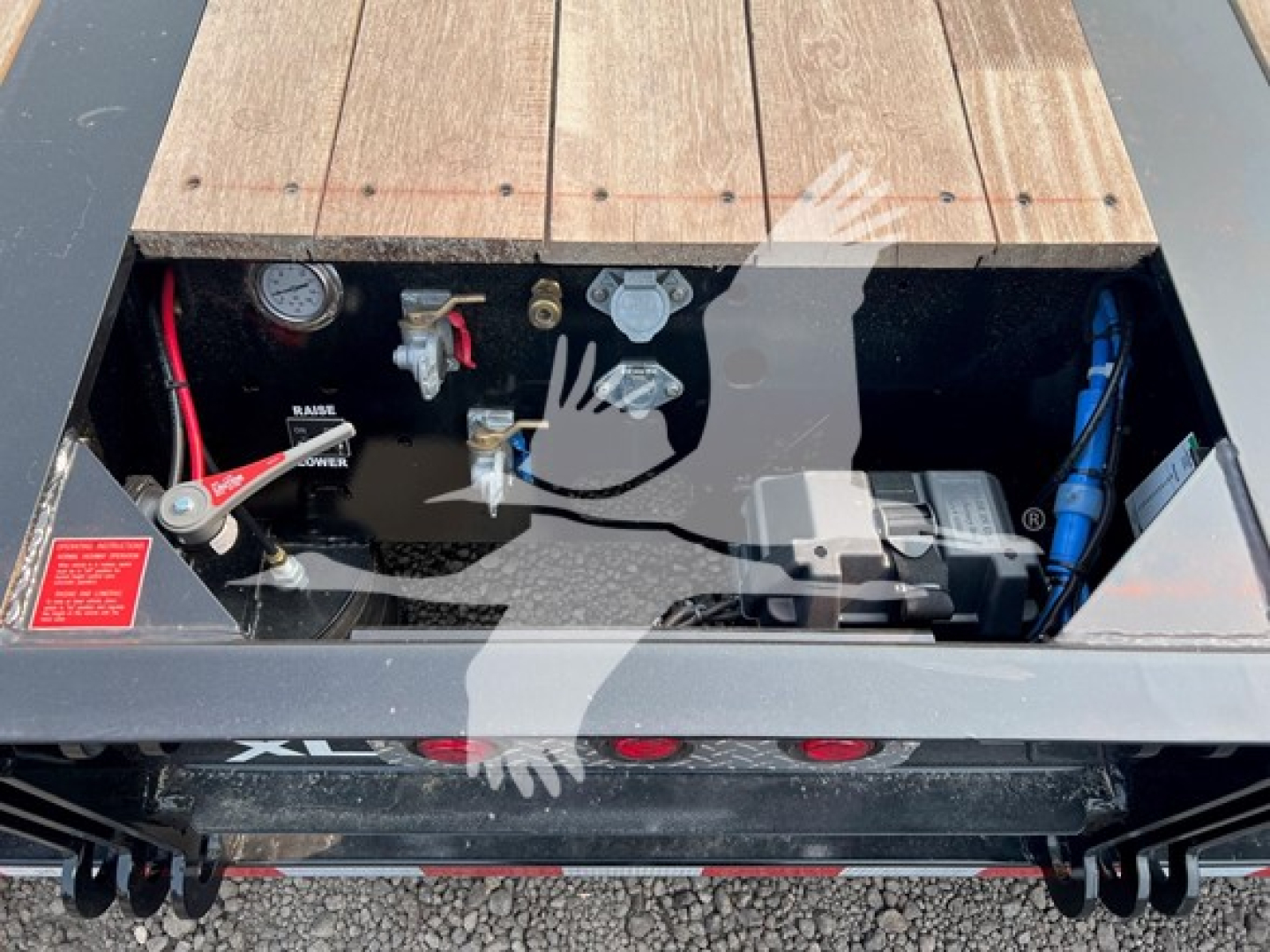 Trailer toolbox area with gauges, hoses, and battery visible under wood deck; hydraulic lines and valves labeled; compact control valve group; red and blue hoses; label stickers.