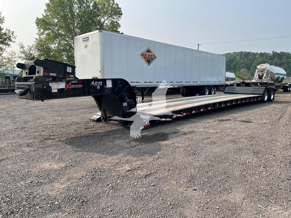 Lowboy trailer with a long flatbed and a white oversized trailer on top, black hitch area, with a diamond placard on the side and a ribbed deck.