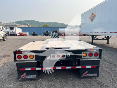 Flatbed trailer rear view showing taillights and mudflaps with XL Specialized Trailers branding; asphalt yard with trailers and a building in background.