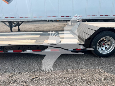 Trailer deck of a lowboy trailer with metal deck and wheel hub visible, side railing and hydraulic components, no text clearly legible.