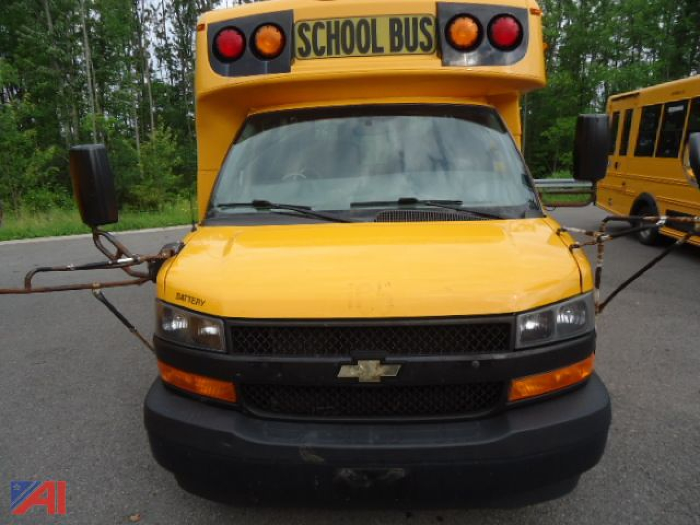 Front view of a 2018 Chevrolet Express micro bus with school bus roof sign, yellow body, black grille, and amber lights, parked with another bus in background.