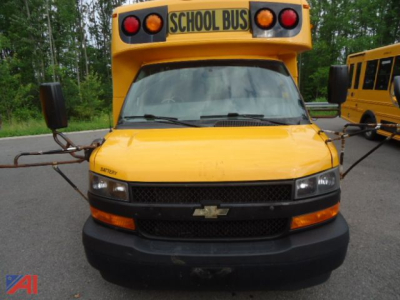 Front view of a 2018 Chevrolet Express micro bus with school bus roof sign, yellow body, black grille, and amber lights, parked with another bus in background.