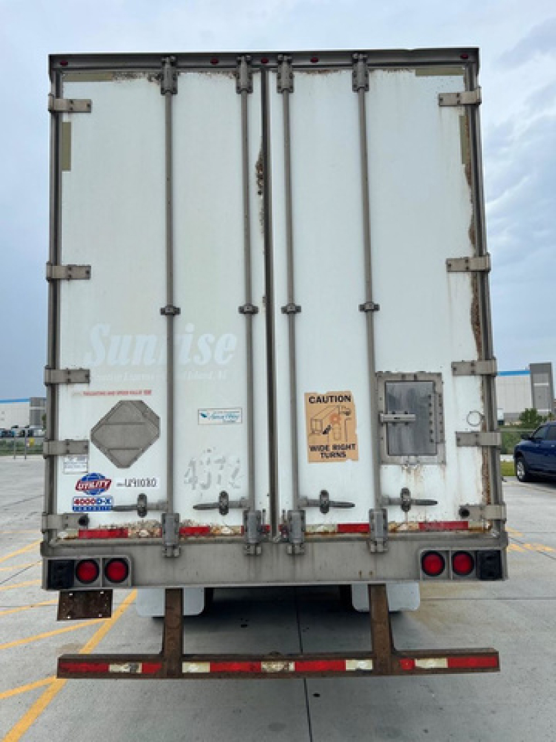 Rear view of a box trailer with double doors showing several stickers and warning labels. Notable text includes 'Sunrise' faintly on the left door, 'CAUTION' label, small sticker with GD? font, and a serial/ID plate near right door. Lights 