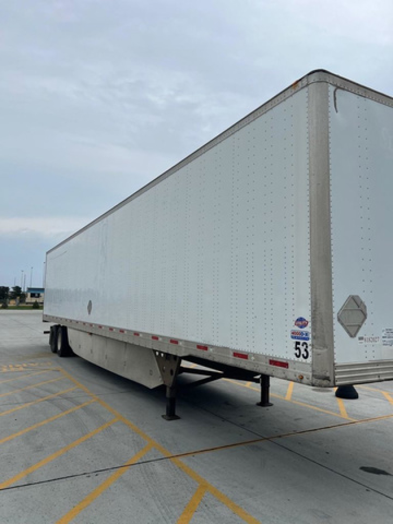 Utility van trailer side view showing a white enclosed trailer with landing legs down and a wheel in the rear, DOT/badge stickers near the front corner.