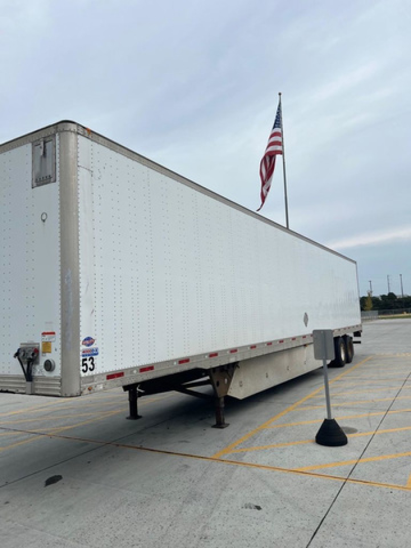 Utility van trailer side view showing white rectangular box trailer with US flag and parking lot lines.