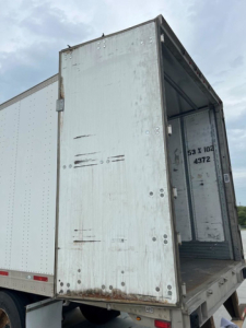 Trailer van interior showing open rear door with metal panels and cargo area; some rust spots and vertical support beams visible.