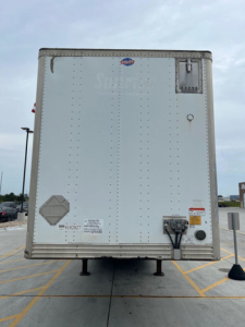 Rear view of a white cargo trailer showing landing gear legs, rear doors, reflectors, and small stickers.
