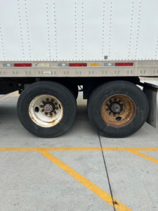 Trailer wheel area of a 2014 utility van showing dual rear wheels with one white rim and one rusted rim, above the wheel well with riveted cargo trailer side.