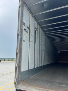 Interior of a cargo trailer showing empty metal walls and floor with open rear doors; inside cargo area visible with support ribs along the ceiling.