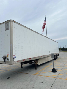 Utility van trailer side view showing white rectangular box trailer with US flag and parking lot lines.