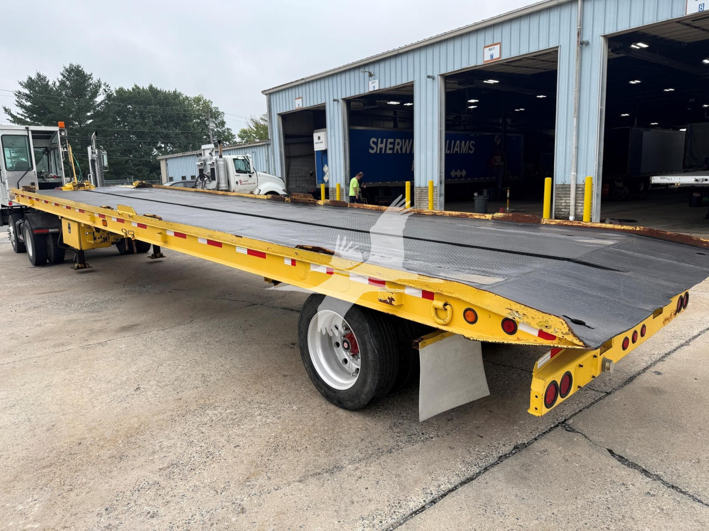 1998 Landoll 330D-1 trailer with long flatbed and damaged non-slip deck. View shows yellow trailer frame, black deck, rear lights, and a wheel. In background, a blue warehouse and other equipment.