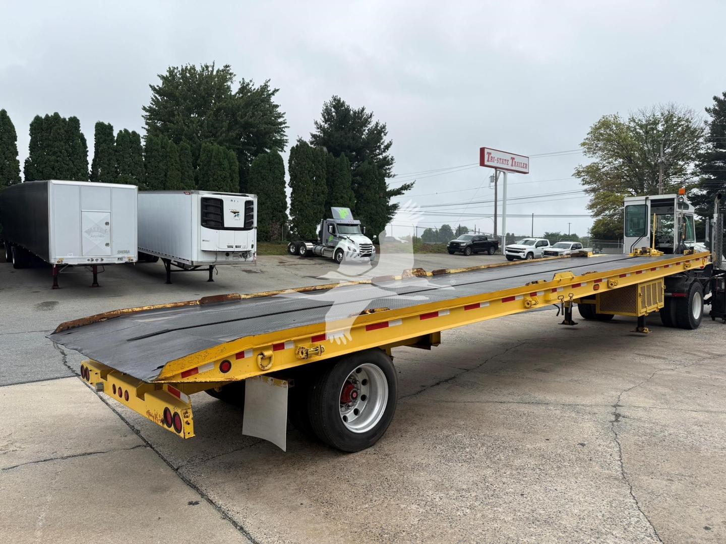 Landoll 330D-1 trailer in yard with long flatbed and gooseneck, showing front hitch and rear dual axles; yellow trailer with black deck, surrounding equipment in background.