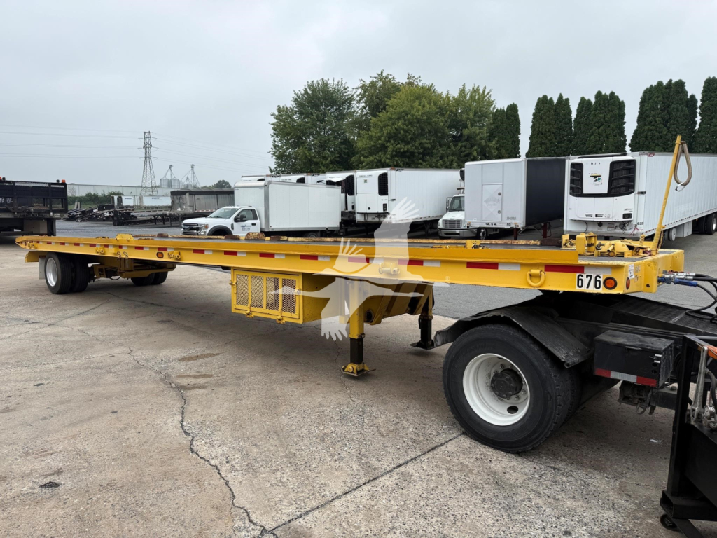 Truck trailer showing a long yellow flatbed trailer with a landing gear and dual rear axles; side shows numbers and equipment; background includes other trailers and a white pickup.