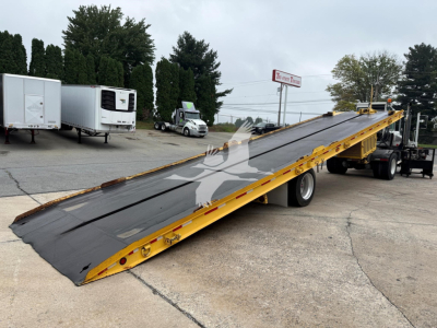 A long yellow Landoll tilt bed trailer with a black surface, parked on a lot. Visible text includes 'Landoll' on the side and a small safety decals along the edge. The scene shows other trailers in background.