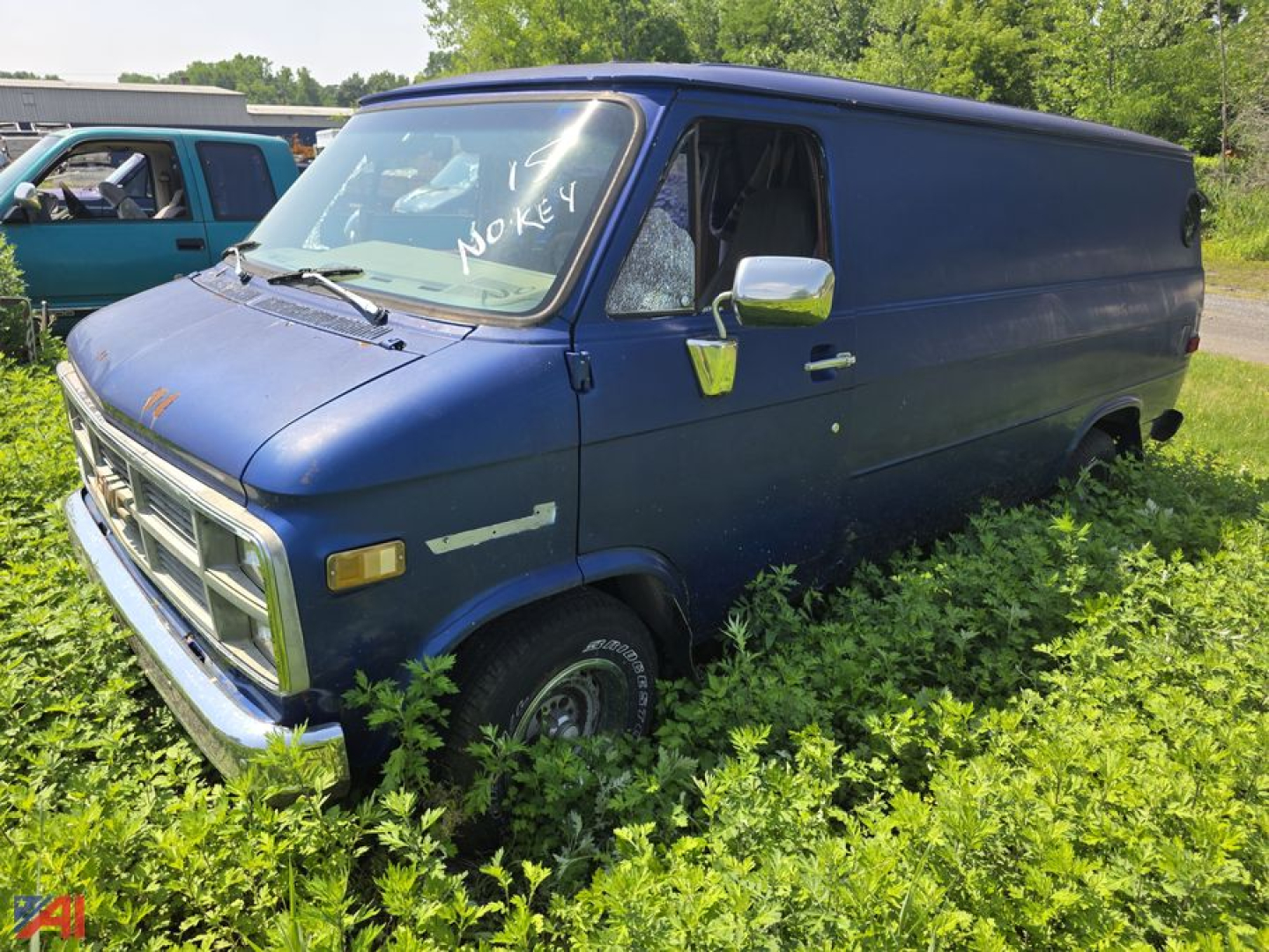 1984 GMC Vandura full-size cargo van in blue, side view parked in tall grass with overgrown weeds, sign on windshield reading 'NO KEY'