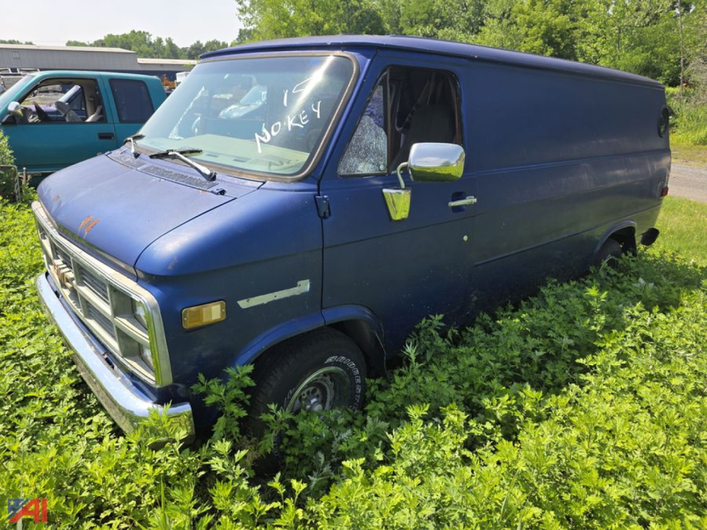 1984 GMC Vandura full-size cargo van in blue, side view parked in tall grass with overgrown weeds, sign on windshield reading 'NO KEY'