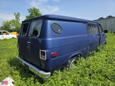 1980s GMC Vandura cargo van in blue with broken rear window, circular porthole window on side, overgrown grass around tires, rear bumper chrome, faded paint.