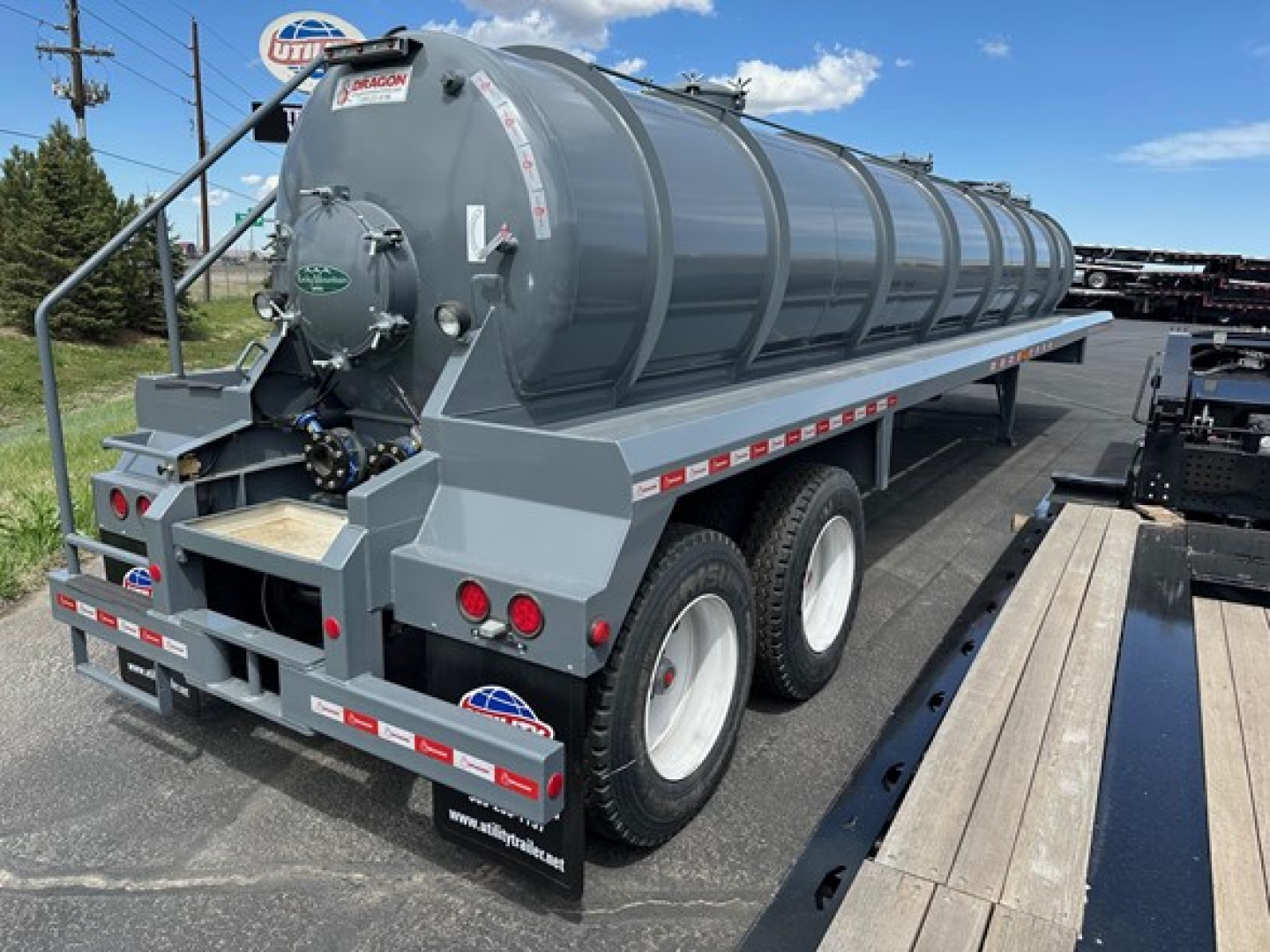 Tank trailer showing a gray vacuum tank with rear access hatch and dual rear axles, side rails, and reflective tape.