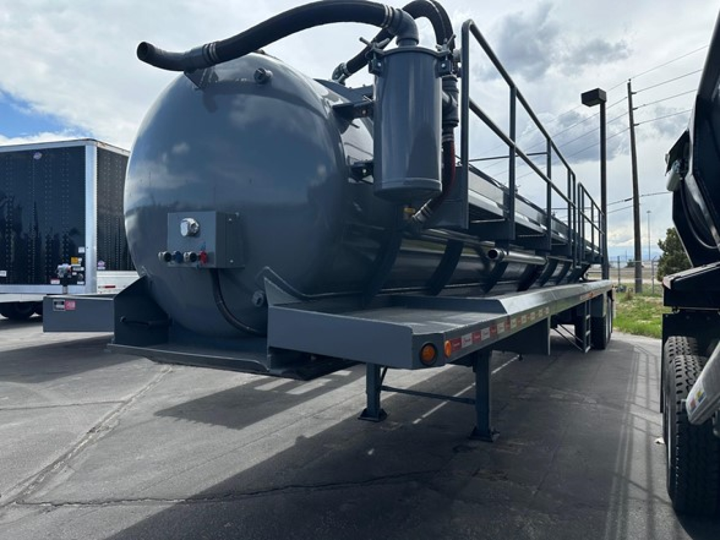 Tank trailer on a flatbed trailer with vacuum system, cylindrical tank with hoses and piping, support legs, and a control panel near the front.