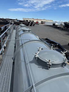 Vacuum trailer tank on a Dragon 130 Bbl storage unit with manway hatch and securing latches running along top of cylindrical tank; industrial yard in background.