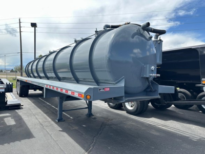 Horizontal cylindrical vacuum tank trailer on a drop- deck trailer with gray finish, round end cap, hoses and fittings, side rails, and support legs visible.