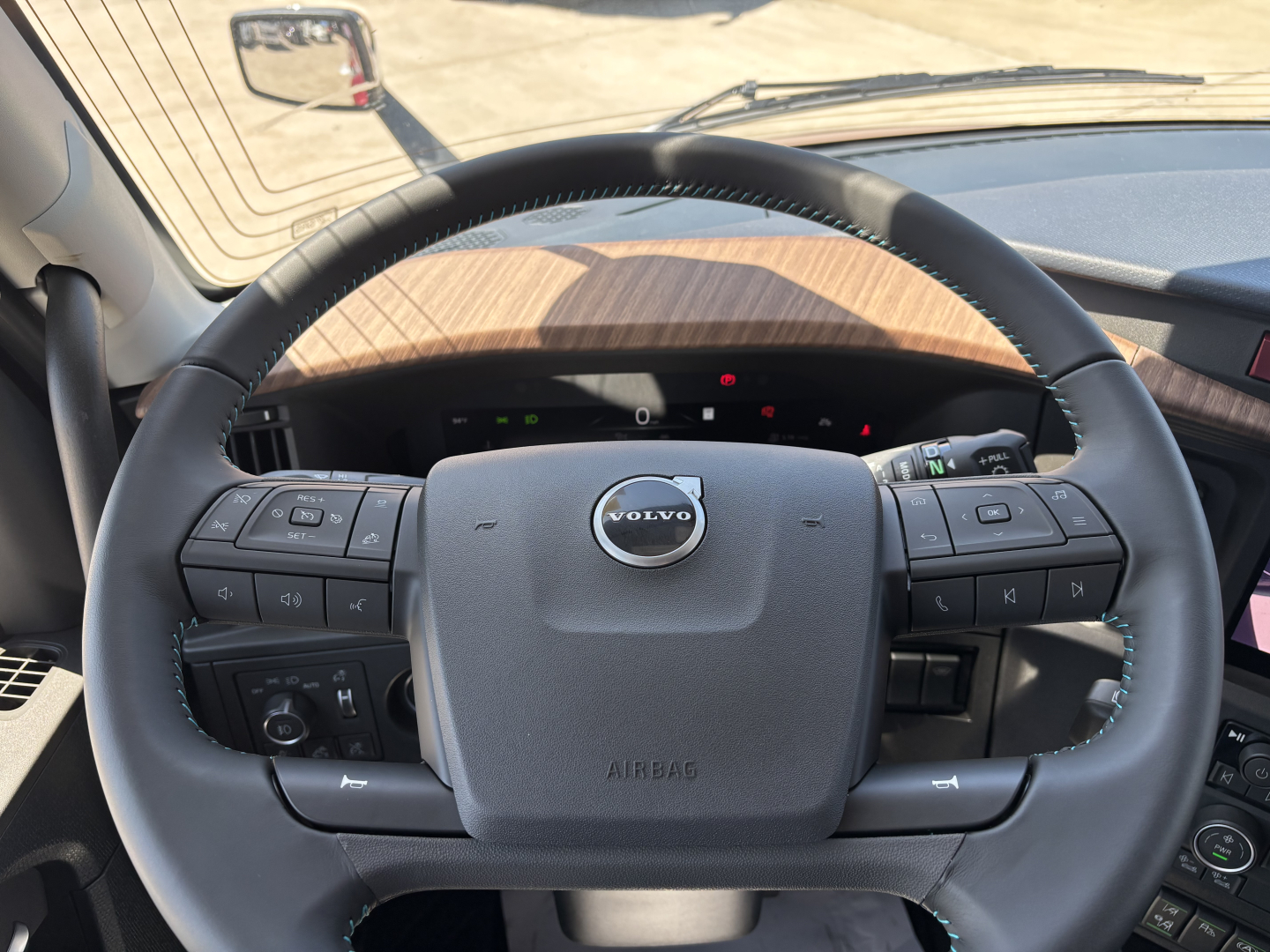 Volvo steering wheel and dashboard view from driver seat of 2027 Volvo VNL truck showing steering wheel with Volvo logo and wood grain dashboard.