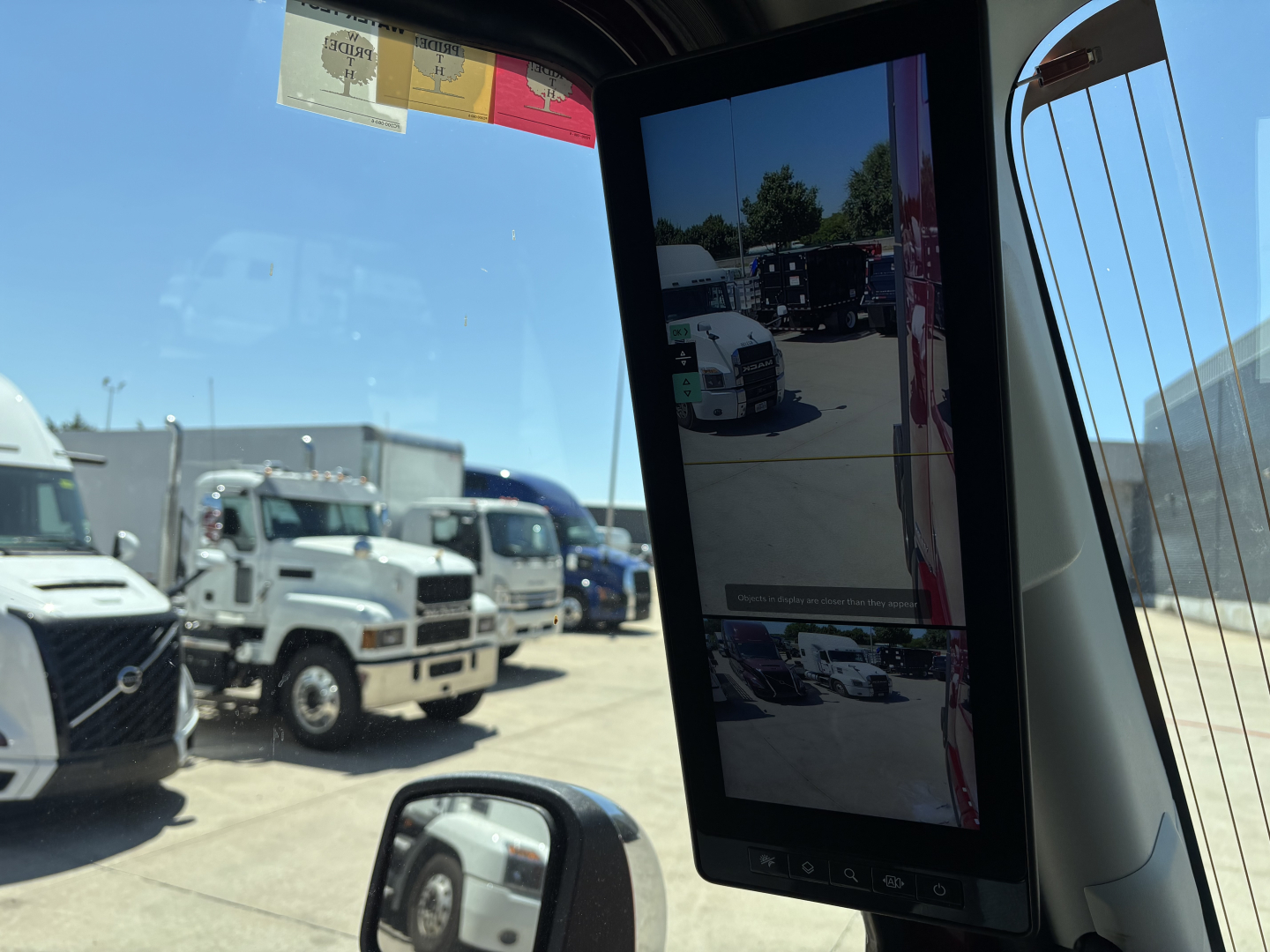 Inside view of Volvo VNL class 8 truck cab with side window, mirrors, and a vertical infotainment screen visible, showroom lot with other trucks outside.